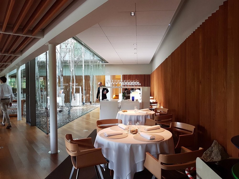 Comedor con manteles blancos, pared de lamas de madera y patio acristalado con abedules